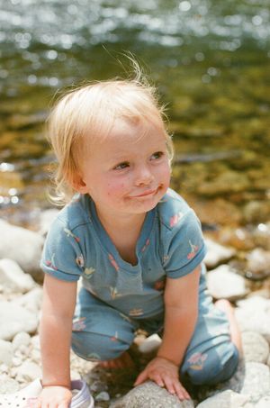 Blonde toddler in blue outfit smiling while playing on rocky riverbank in bright sunlight.