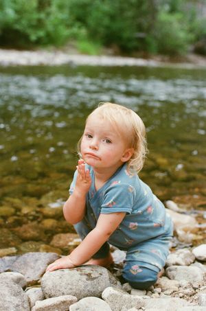 Blonde toddler in blue outfit kneeling on rocky riverbank with hands clasped, looking up curiously.