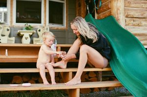 A mother applies sunscreen to her toddler on wooden steps beside a green slide outside a log cabin.