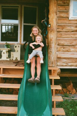 A smiling woman slides down a green playground slide with a toddler on her lap beside a rustic log cabin.