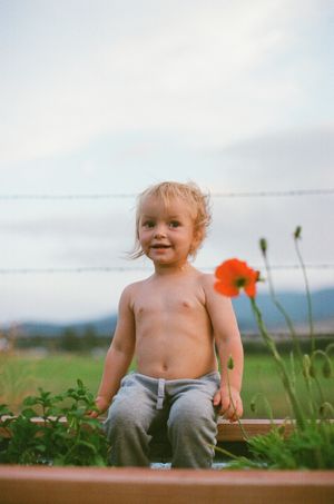 Shirtless toddler sitting outdoors near a red poppy flower with a green field and cloudy sky in the background.