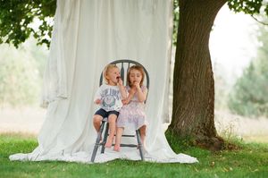Two young children sit on a wooden chair outdoors against a white fabric backdrop near a large tree.
