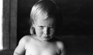 Black and white portrait photo of a young blonde toddler with a serious expression, shirtless, in soft natural light.