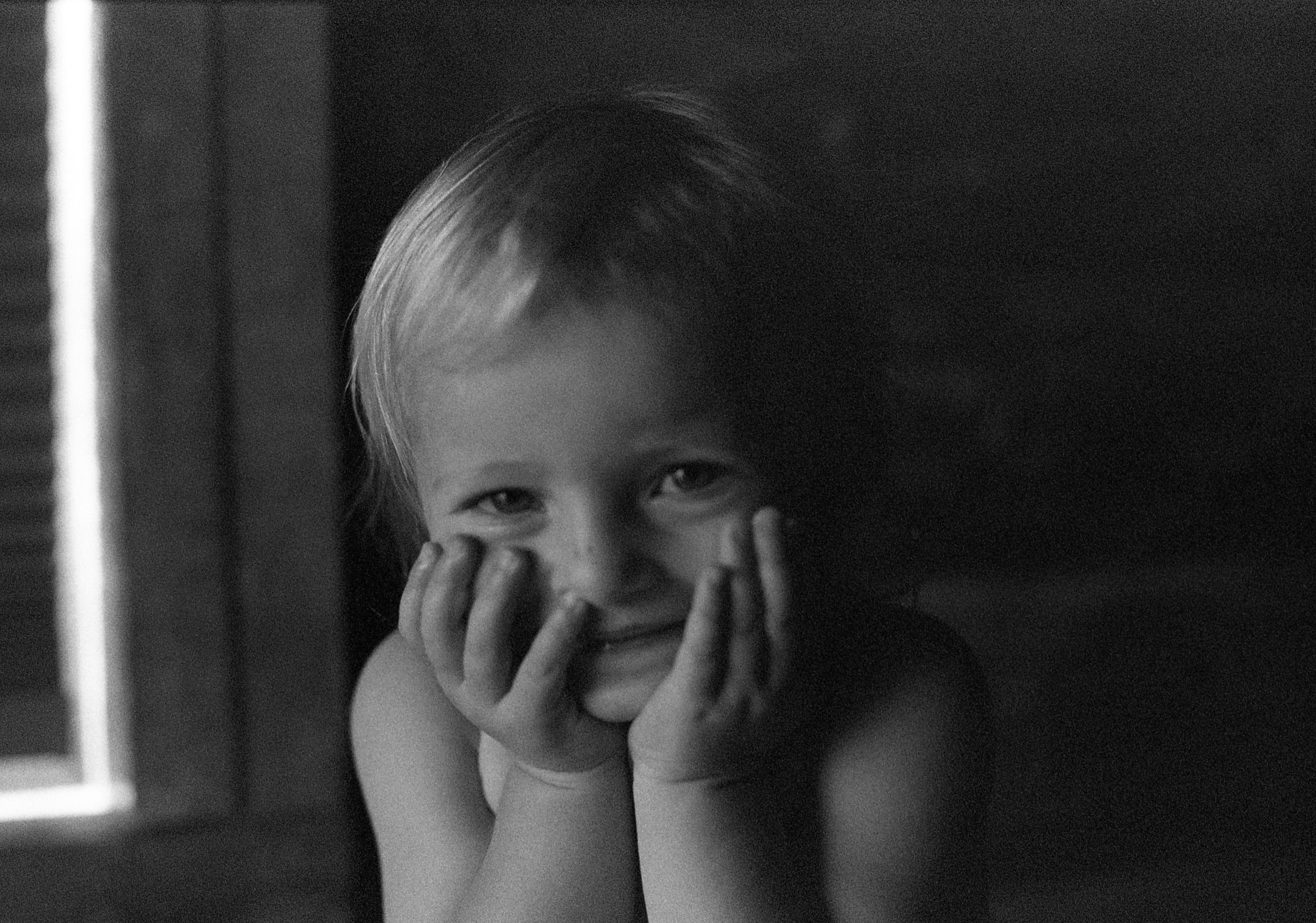A smiling toddler with blonde hair rests chin on hands in a charming black and white portrait photo.
