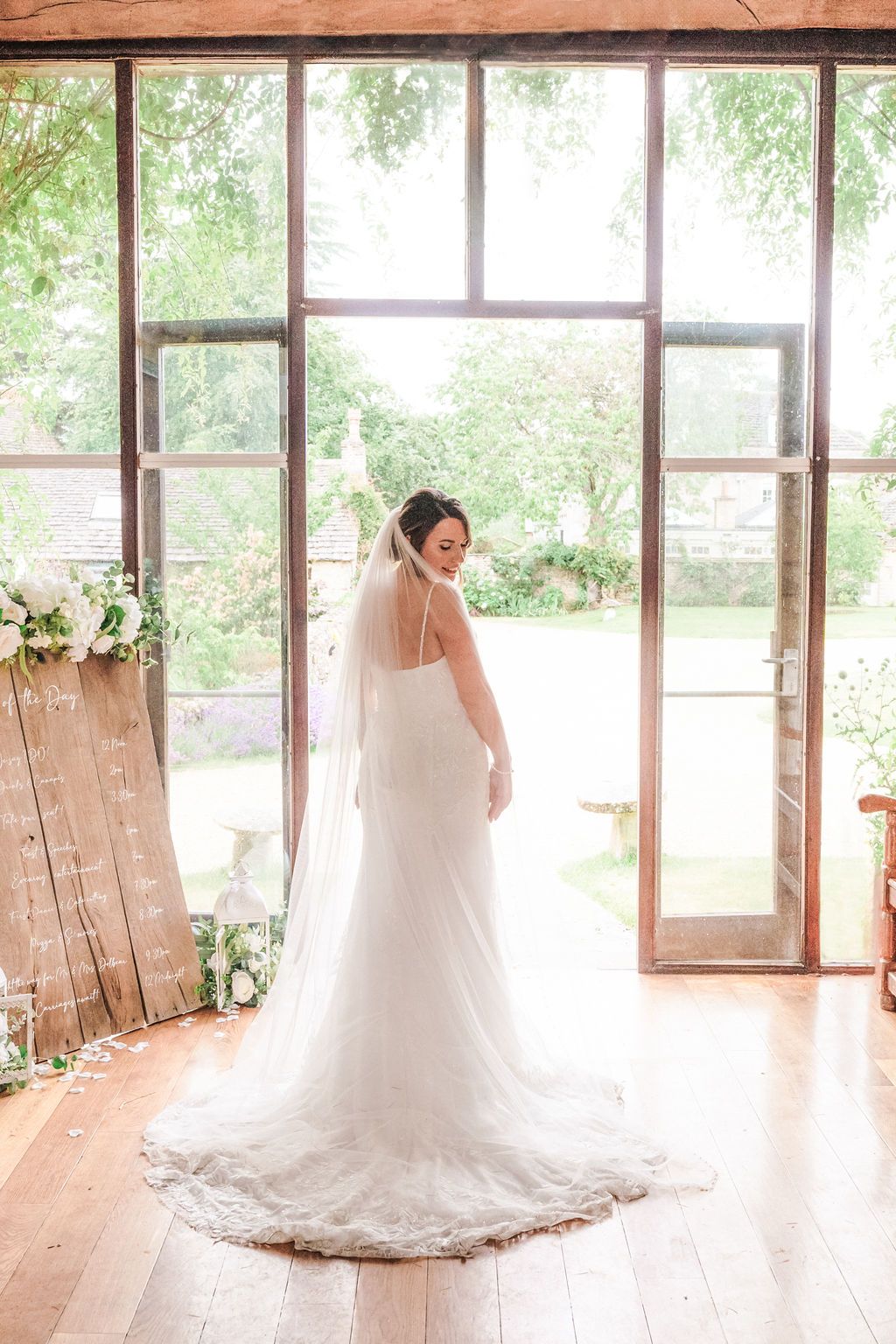 Bride in elegant white lace wedding gown with cathedral veil stands before large glass doors in a sunlit venue.