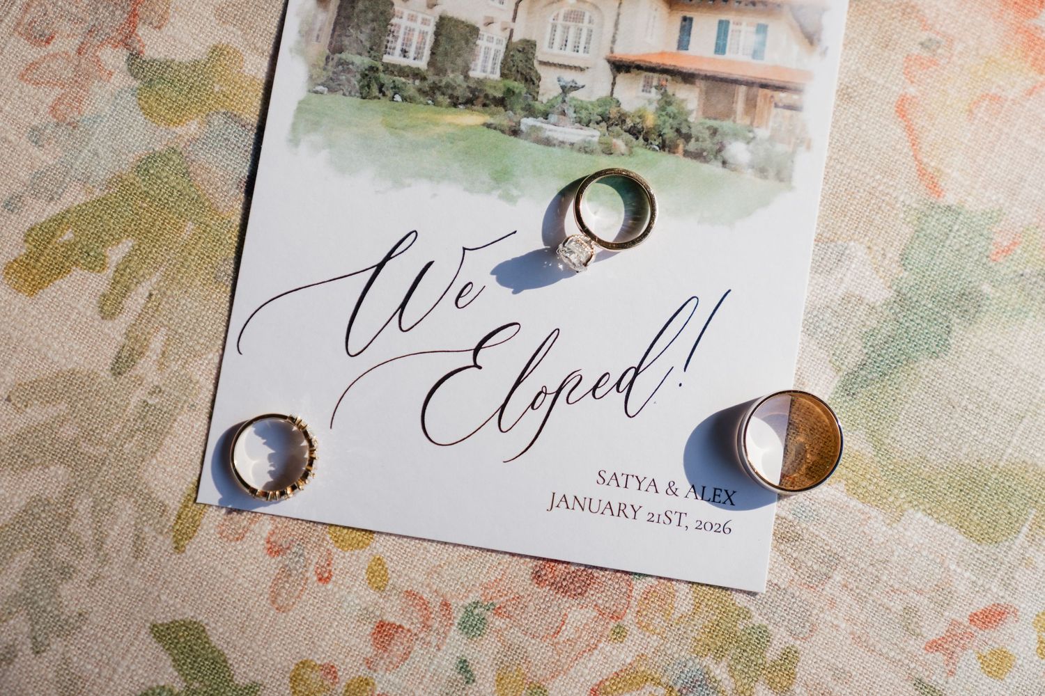 Two gold wedding bands rest on a watercolor elopement announcement card reading We Eloped with a venue illustration.