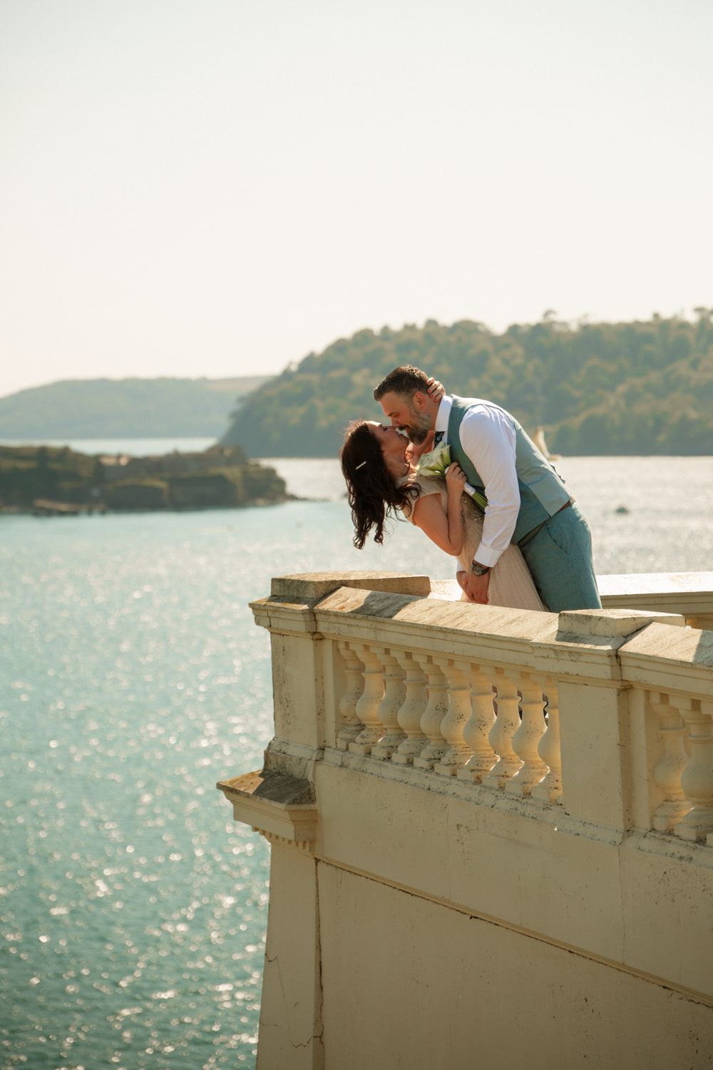 A couple shares a romantic kiss on a stone balcony overlooking a shimmering turquoise sea and lush green island.