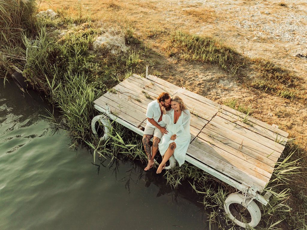Couple sitting together on a rustic wooden dock by a calm river surrounded by lush green reeds.