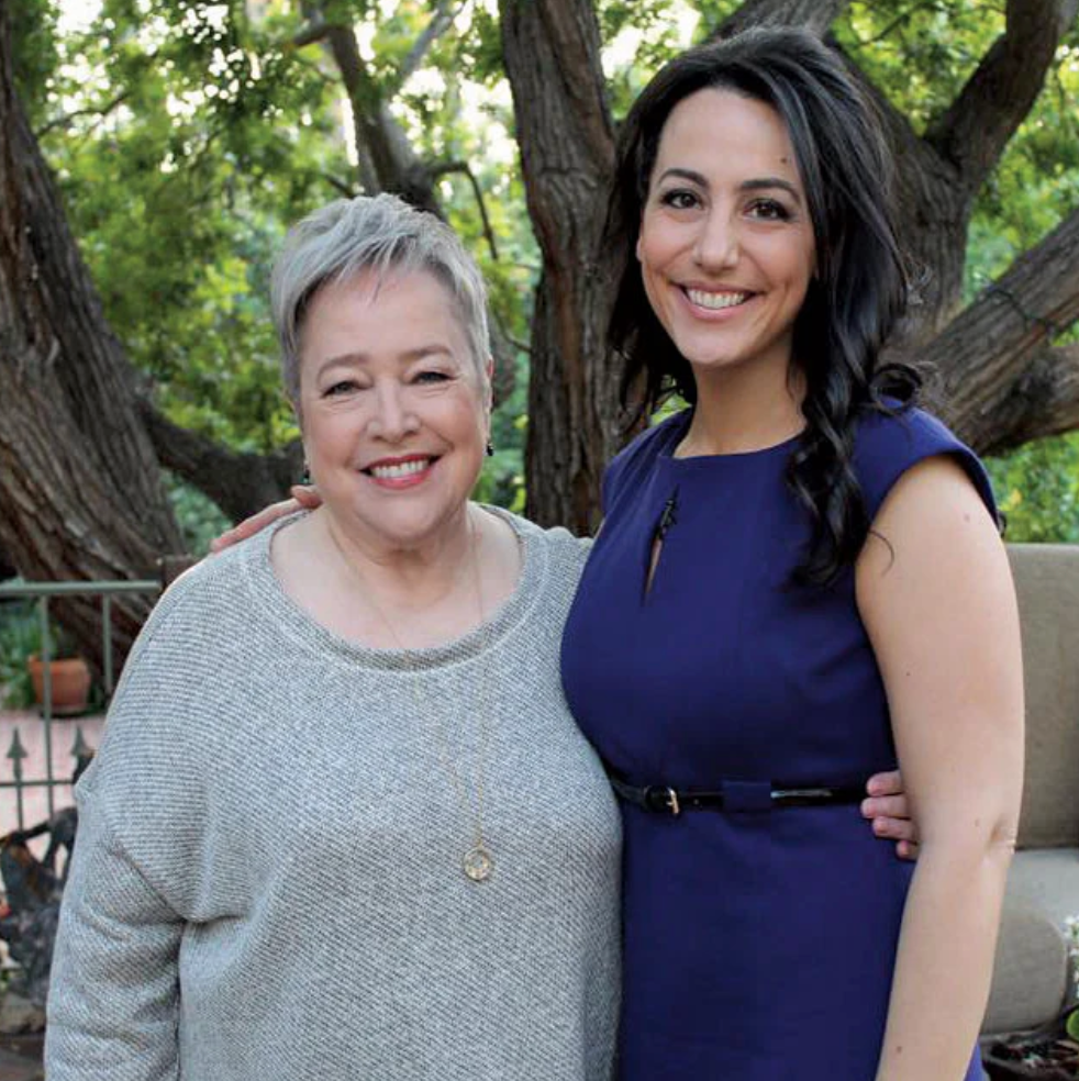 Two women smiling together outdoors, one in a gray sweater and one in a blue dress, posing in front of large trees.