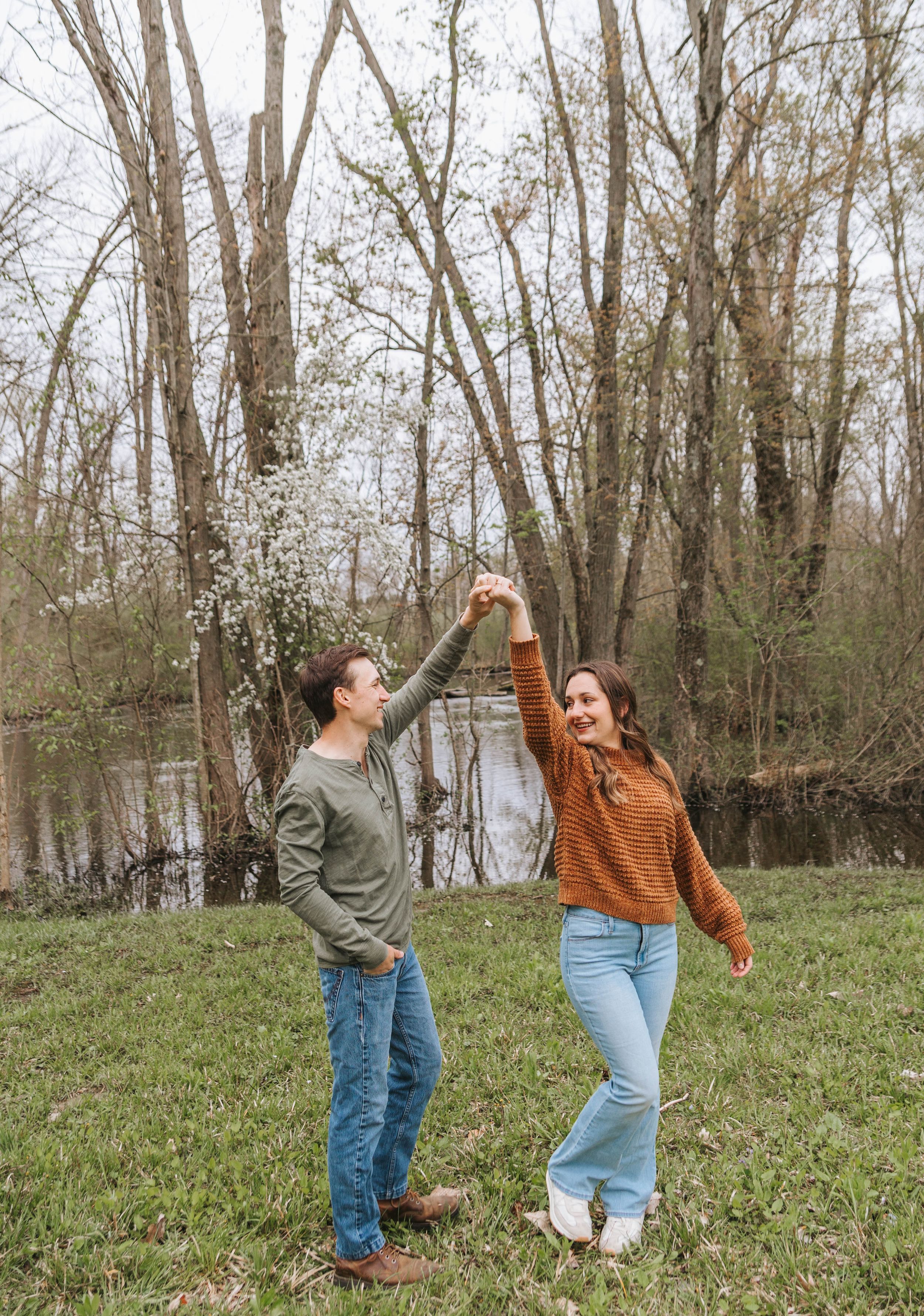 A couple dances outdoors near a pond, holding hands raised, surrounded by bare trees in early spring.