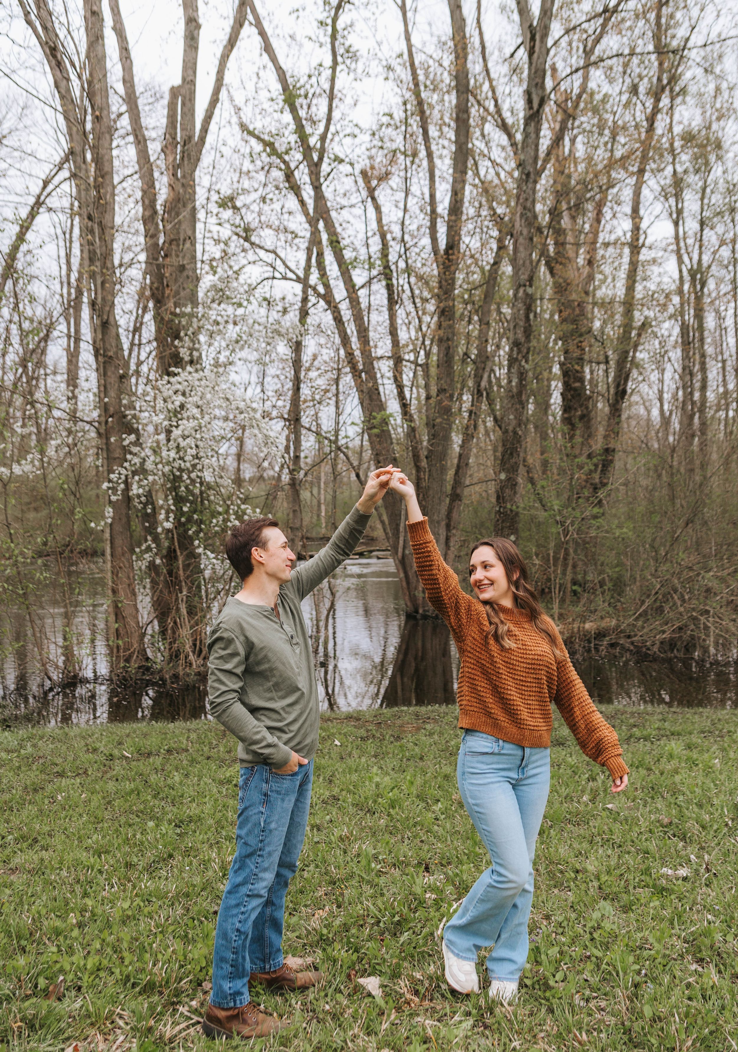 A couple dances outdoors near a pond, surrounded by bare trees in early spring, as the man spins the woman joyfully.