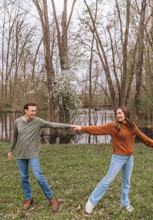A smiling couple holds hands and twirls outdoors near a pond surrounded by bare trees in early spring.
