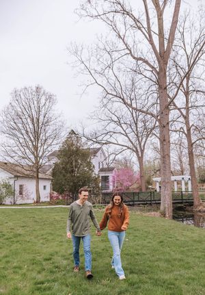 A couple walks hand-in-hand across a green lawn near a white farmhouse and bare trees in early spring.