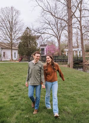 A smiling couple walks hand-in-hand across a green lawn with bare trees and a white farmhouse in the background.