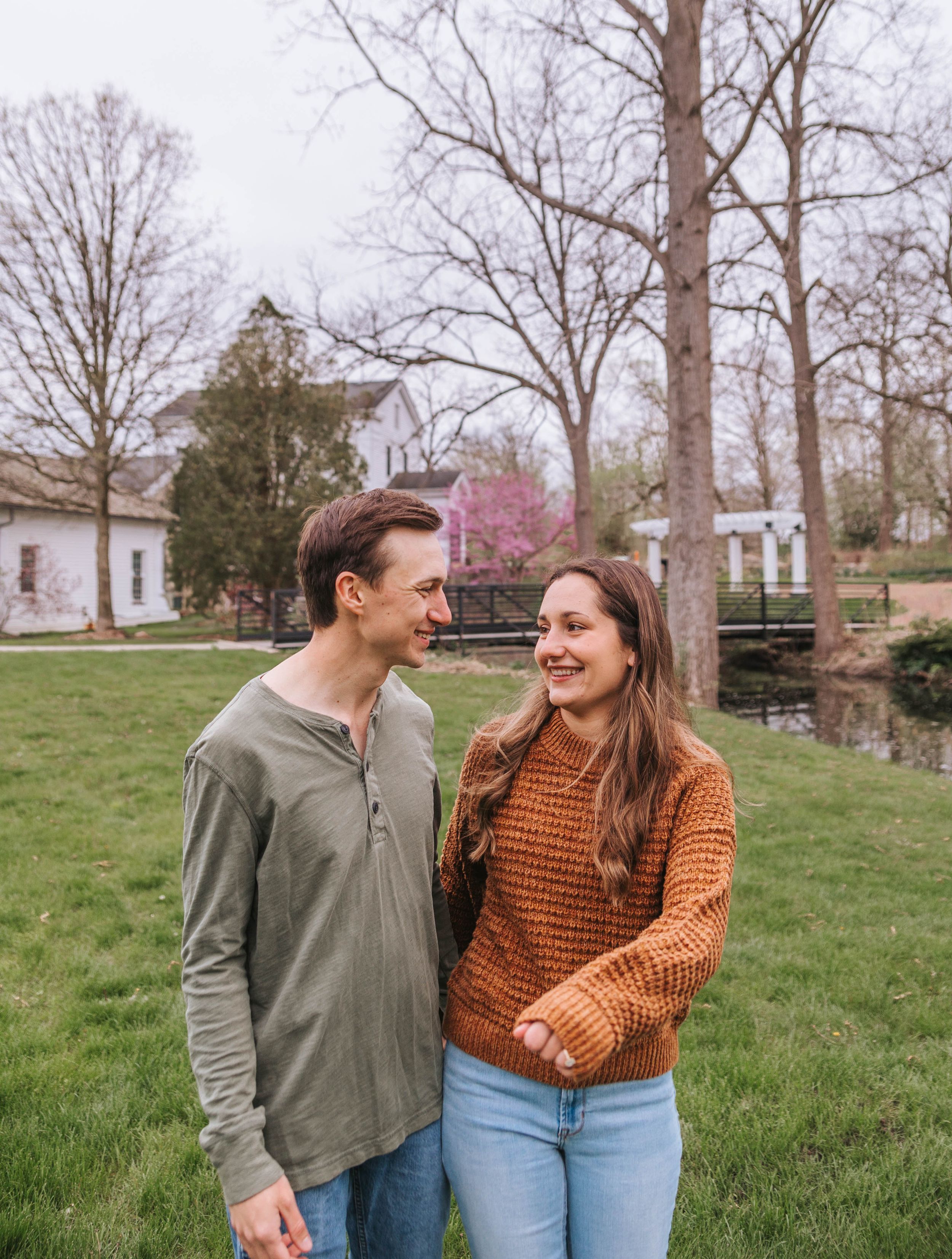 A smiling couple walks together on a green lawn in spring, with bare trees and a white farmhouse in the background.