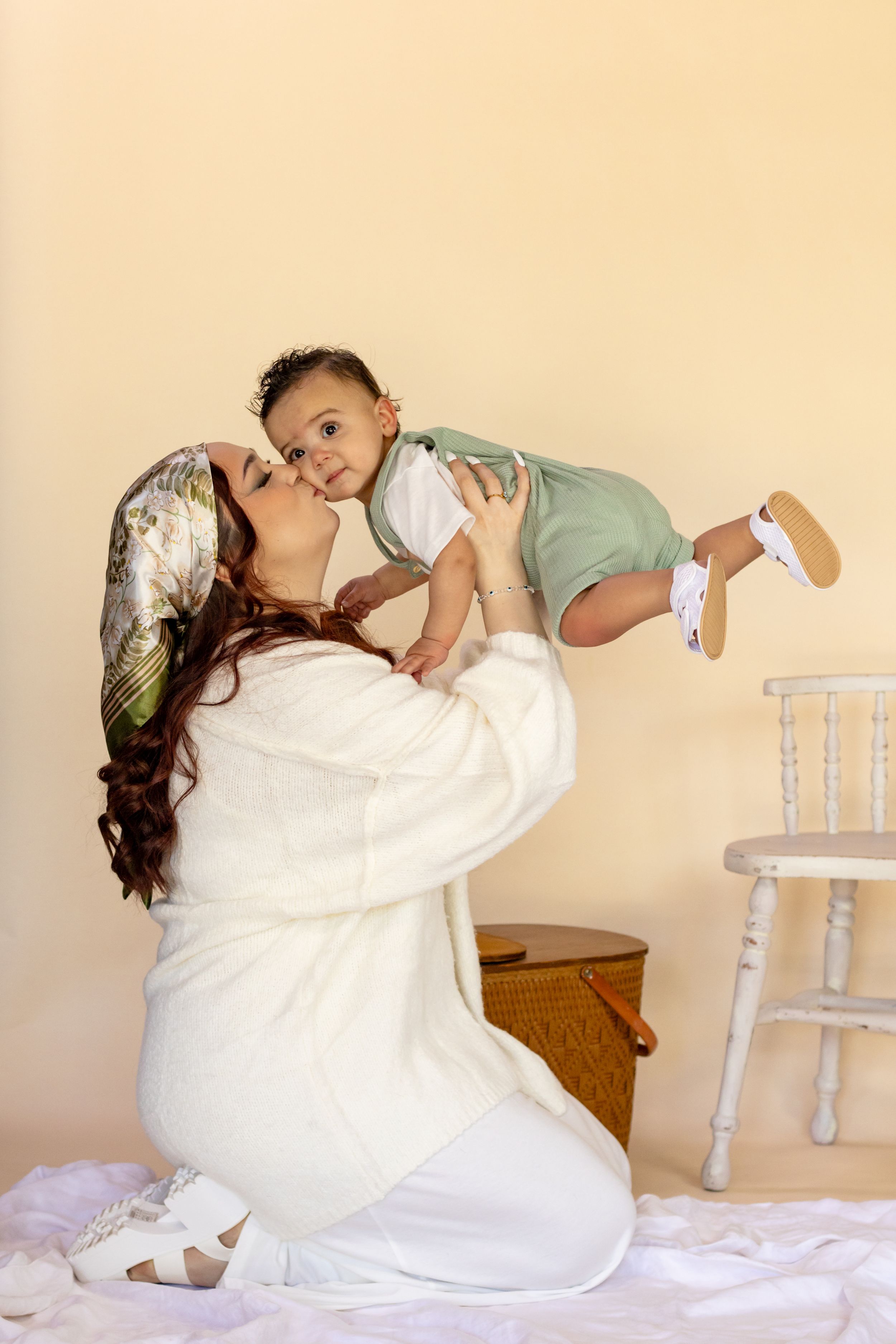 A smiling mother in a white outfit lifting and kissing her baby boy with bright neutral backdrop in roy studio by pierce couny photographer