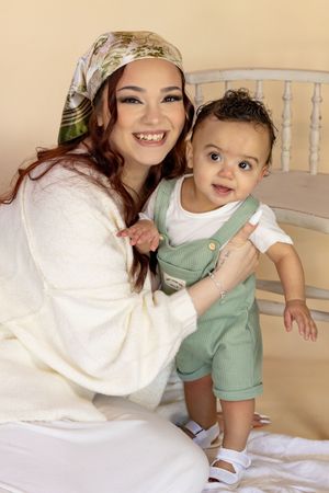 A joyful mother wearing a floral headscarf posing with her adorable baby boy in green overalls taken by JBLM photographer in Roy WA studio.