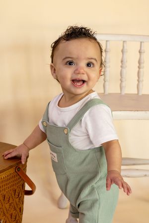 Smiling baby in green overalls and white shirt standing next to a wooden chair in roy washington studio taken by photographer
