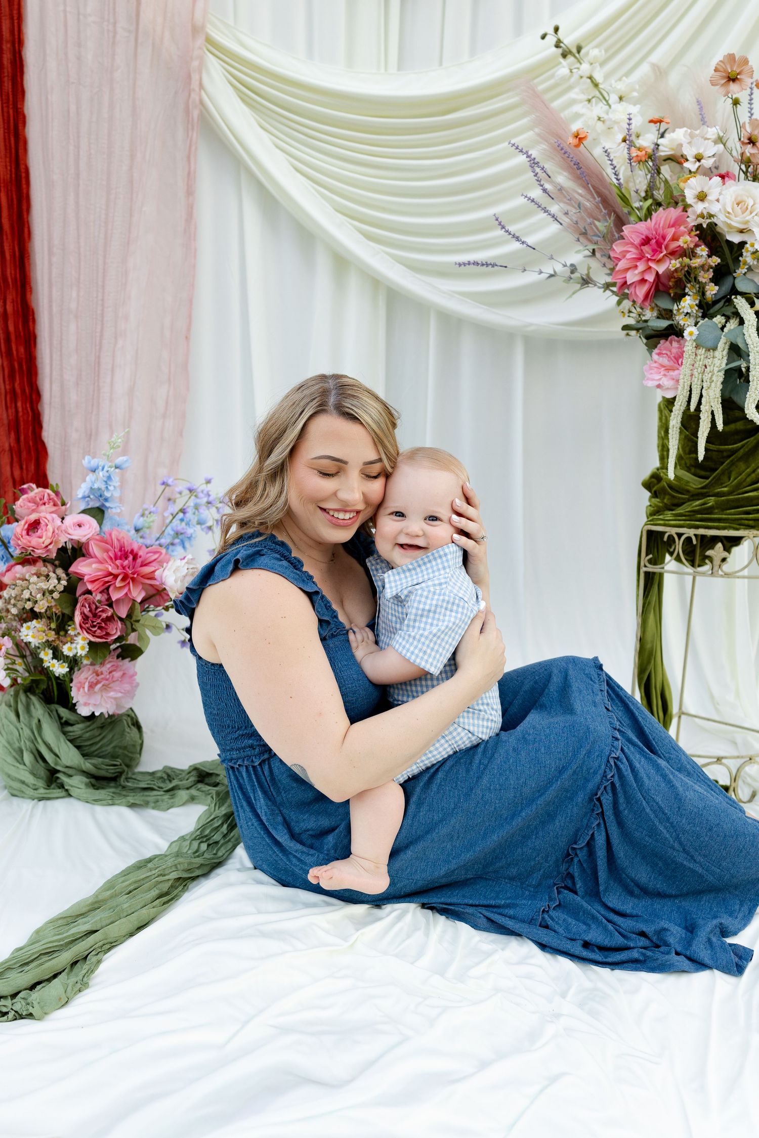 A smiling mother in a denim dress holds her happy baby boy in a floral studio portrait setting.