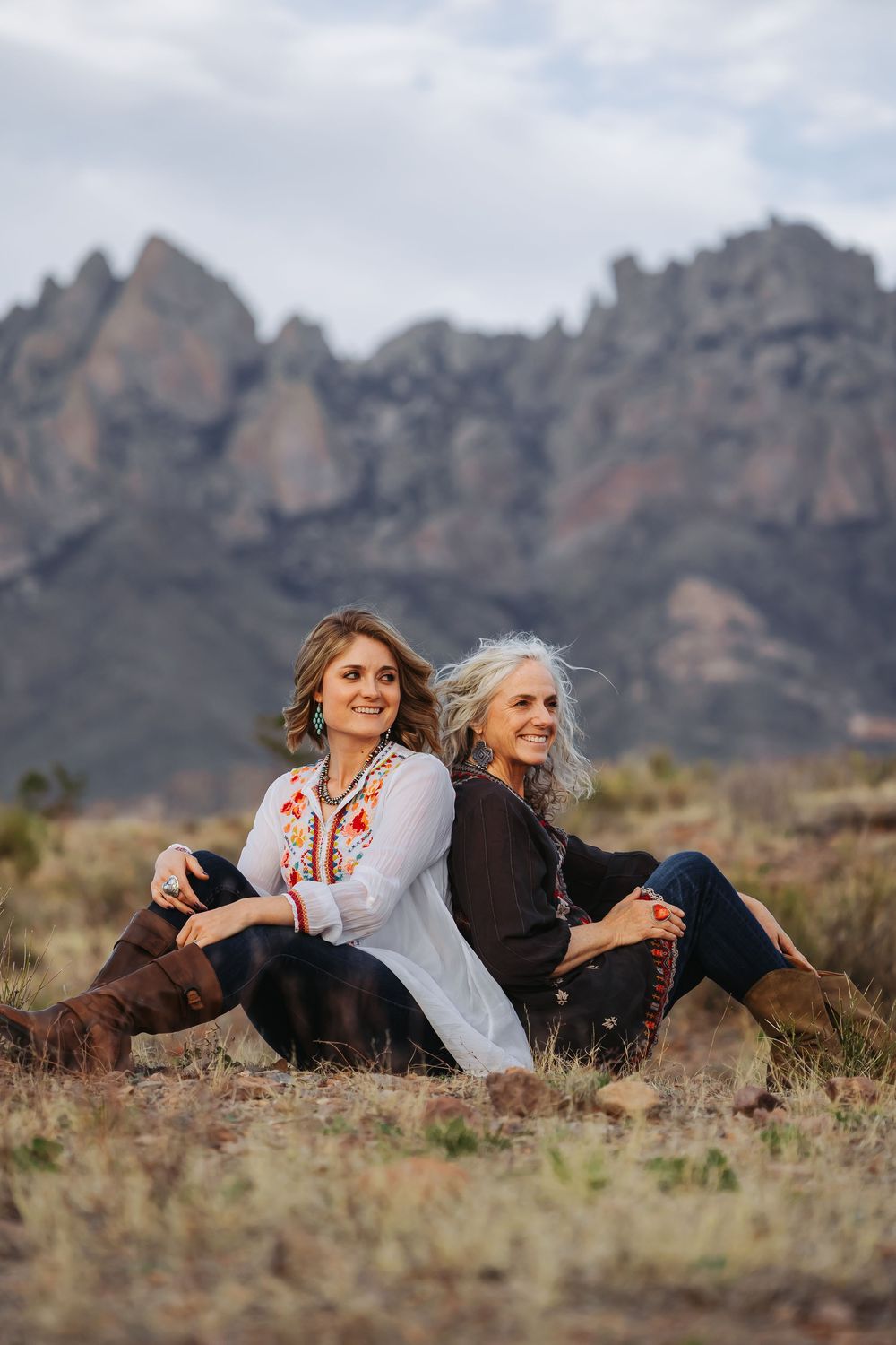 Two women sitting back-to-back in a desert landscape with dramatic rocky mountains in the background.