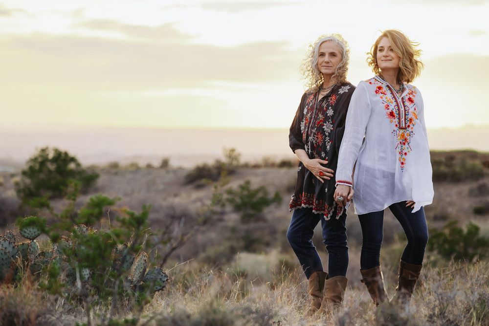 Two women in bohemian floral blouses and jeans pose in a desert landscape at sunset.