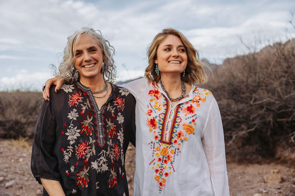 Two women in embroidered bohemian tops smile outdoors in a desert landscape under a cloudy sky.