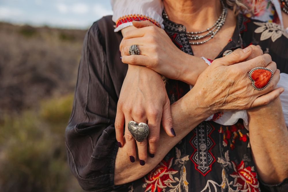 Two women embracing, wearing silver and turquoise rings, red coral bracelets, and boho necklaces outdoors.