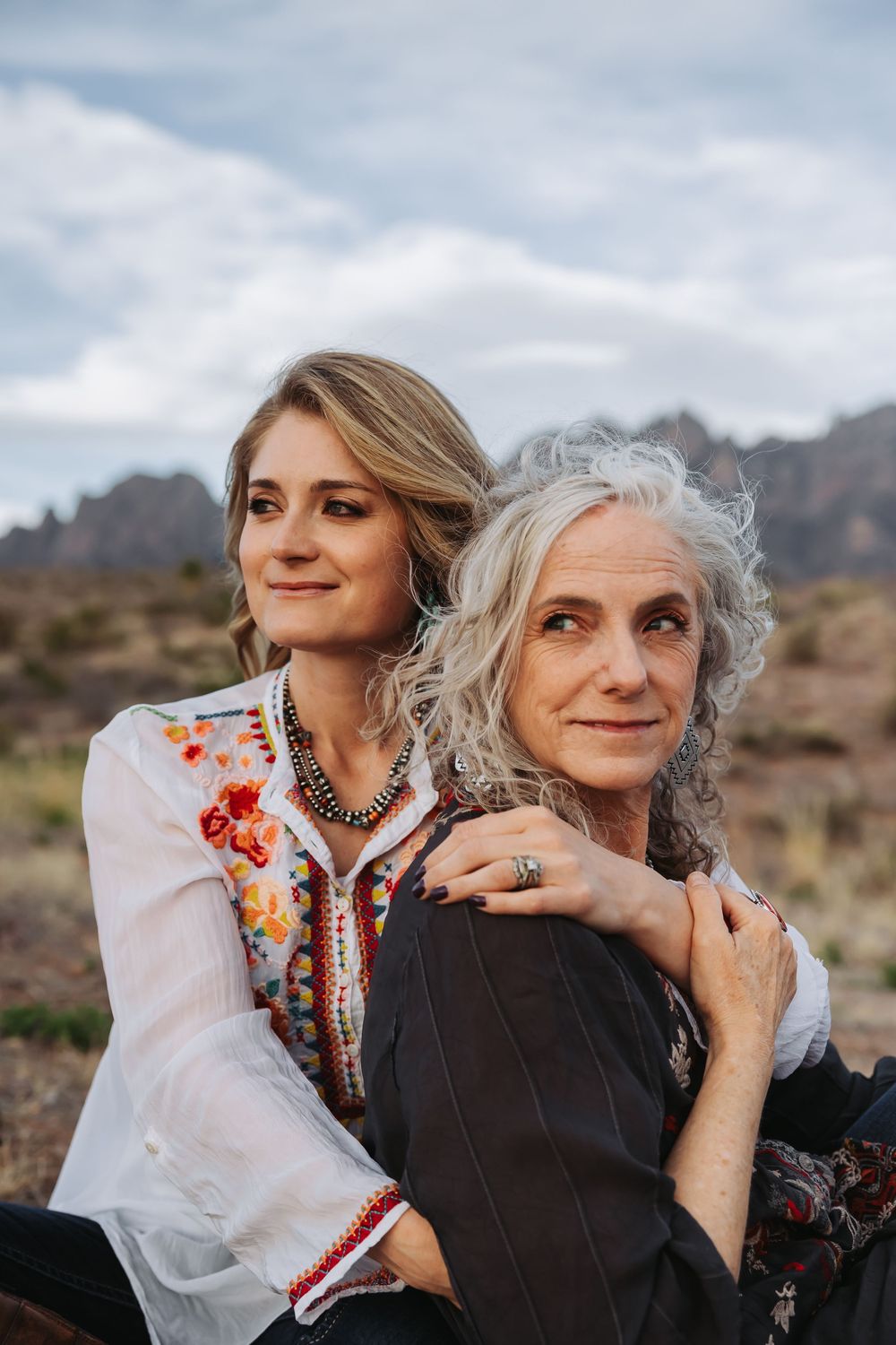 Two women, one young blonde and one older with gray curly hair, pose together outdoors with mountains in the background.