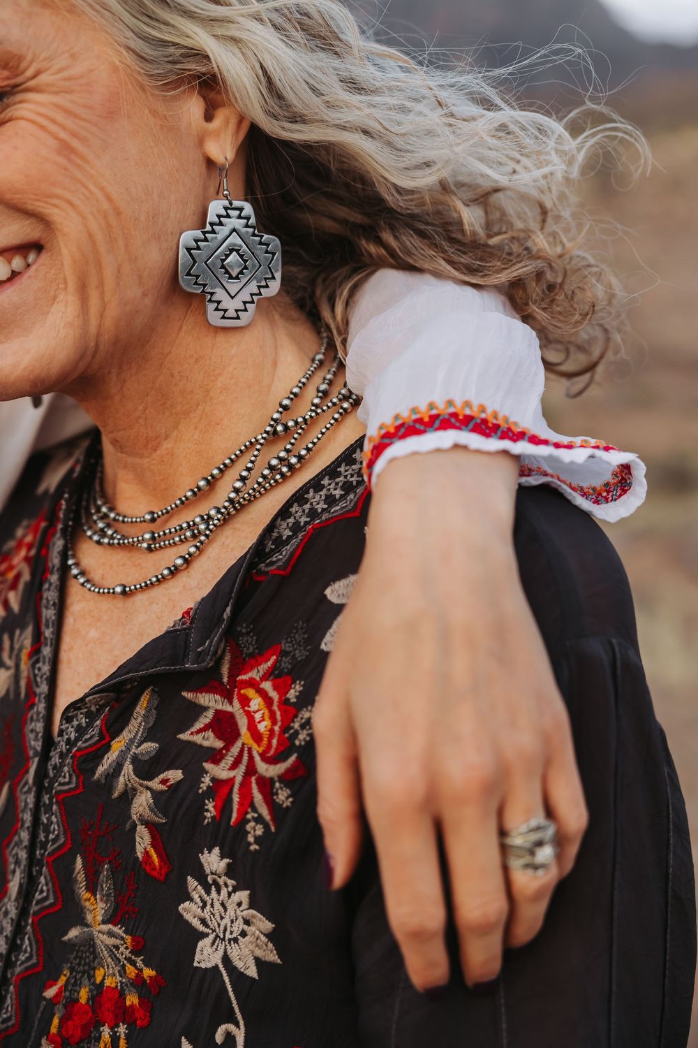 Smiling woman with gray hair wearing Native American-inspired jewelry, floral embroidered jacket, and silver rings outdoors.