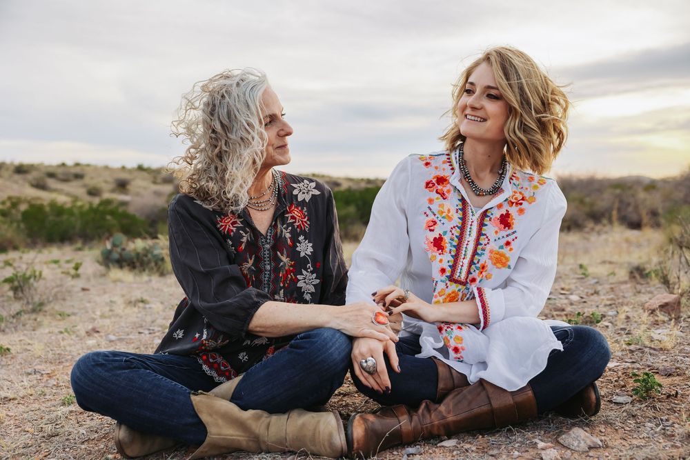 Two women sitting outdoors in a desert field, smiling and wearing floral embroidered tops, sharing a joyful moment.