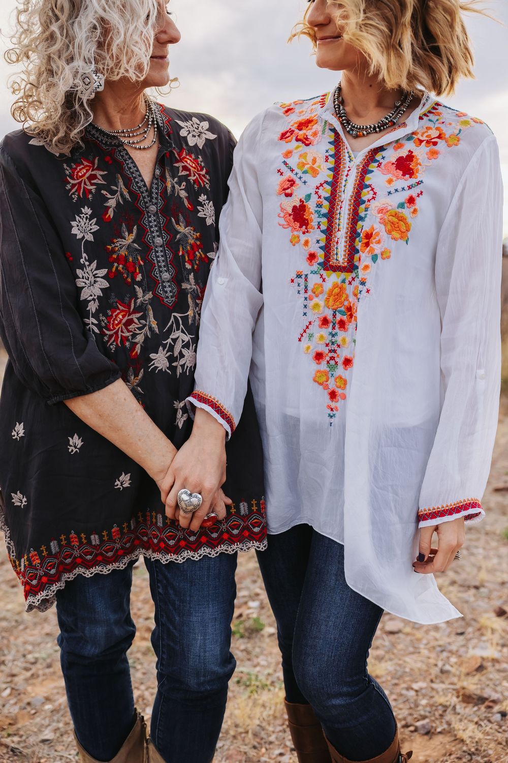 Two women in embroidered boho tunics and jeans hold hands outdoors, one in black and one in white floral top.