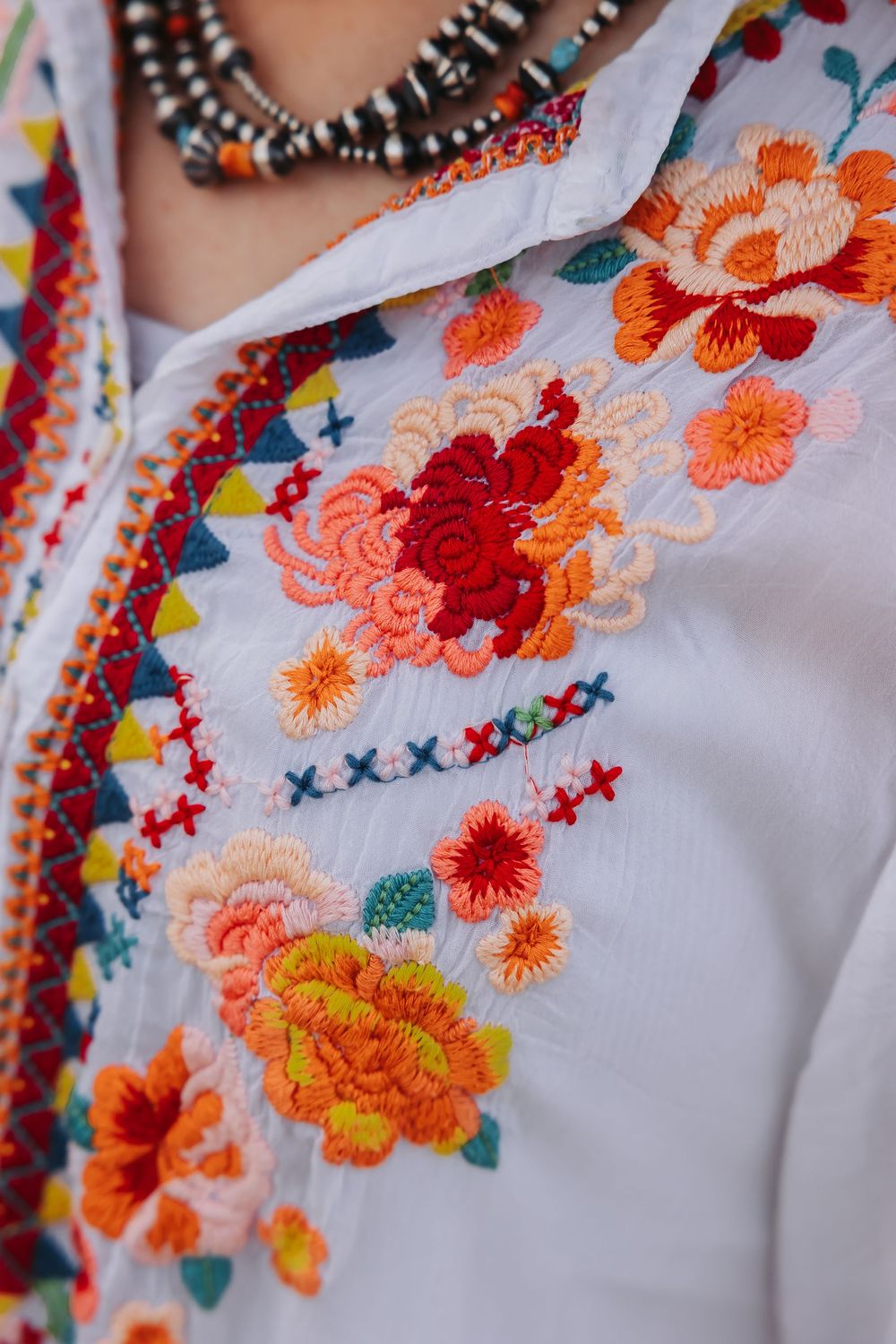 White embroidered blouse with colorful floral and geometric folk patterns, worn with beaded necklace.