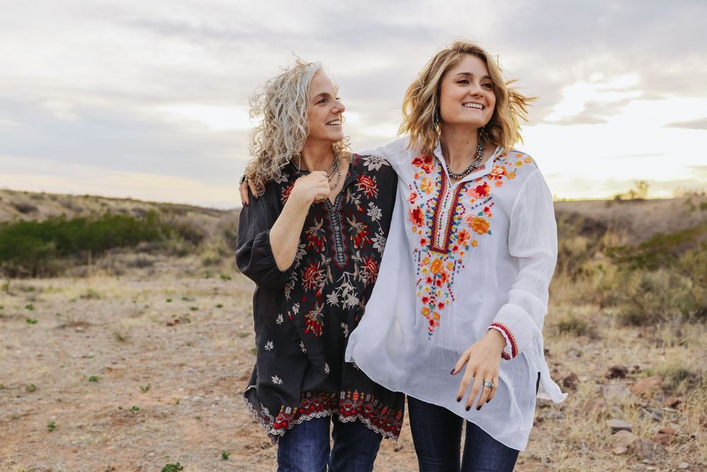 Two women in embroidered boho tops smile and walk together through a desert landscape at sunset.