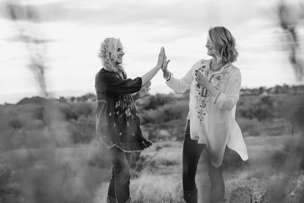 Two women high-fiving outdoors in a desert landscape, captured in black and white photography.