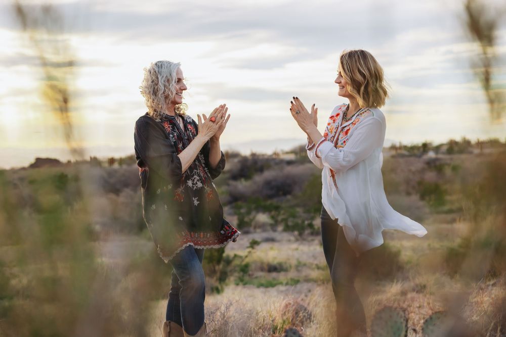 Two women clapping hands together outdoors in a desert landscape at dusk.