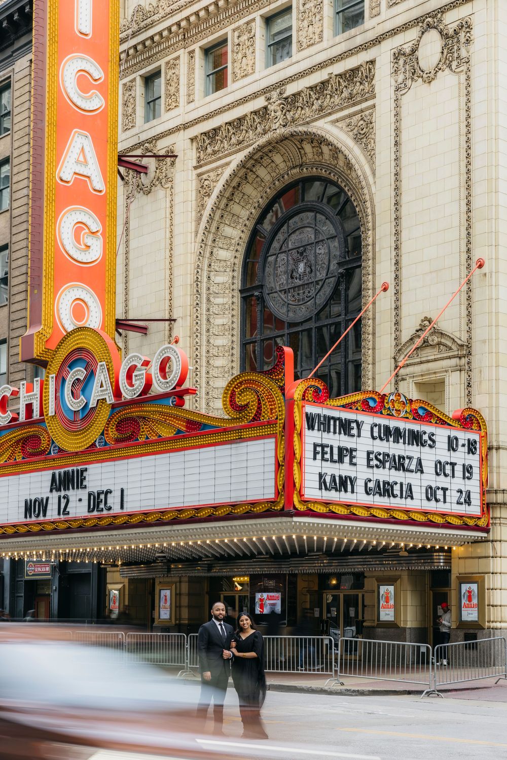 Indian Couple holding hands in front of the iconic Chicago Theatre marquee, dressed in elegant black formal attire during engagement session