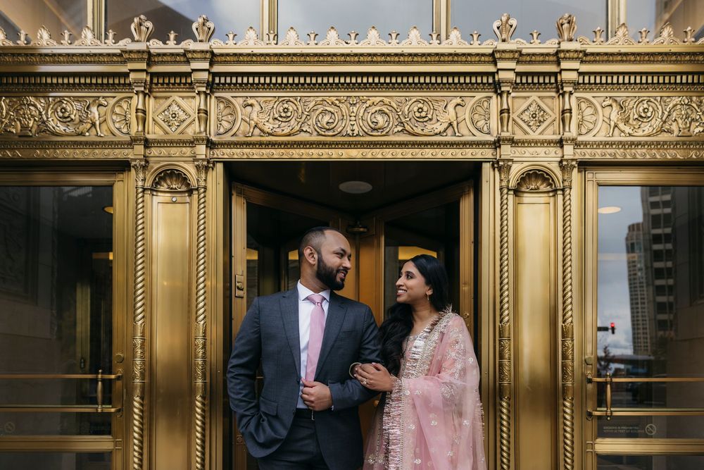 South Asian couple in elegant attire poses lovingly before ornate gold elevator doors by Chicago River Walk during engagement session