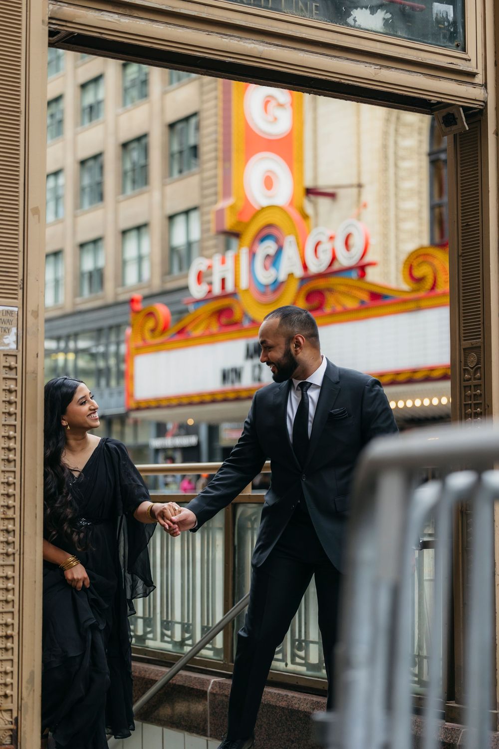 Indian Couple holding hands in front of the iconic Chicago Theatre marquee, dressed in elegant black formal attire during engagement session