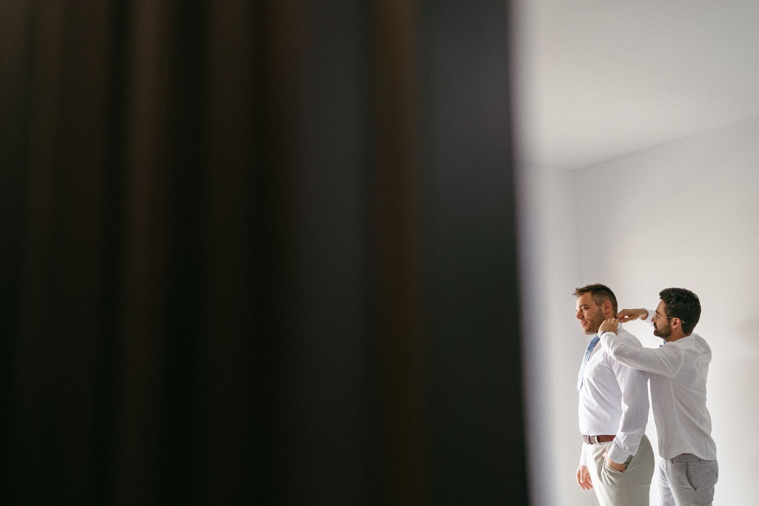 Two groomsmen in white shirts help each other prepare for a wedding ceremony in a bright modern room.