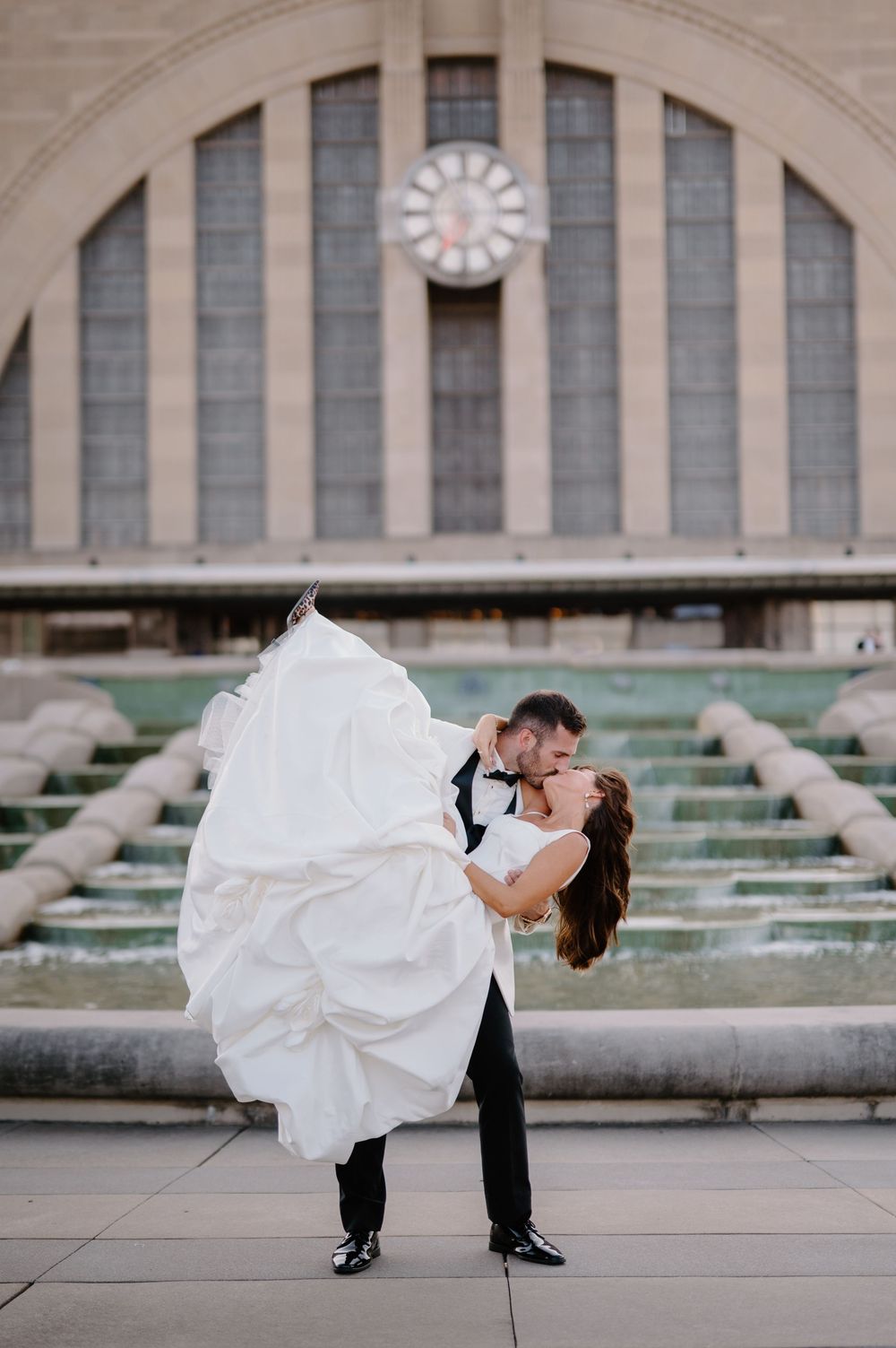 Groom lifts bride in white ballgown for romantic kiss outside Cincinnati Union Terminal fountain.