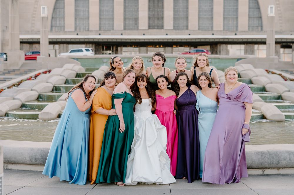 Bride in white gown poses with colorful bridesmaids in mismatched dresses by a fountain.