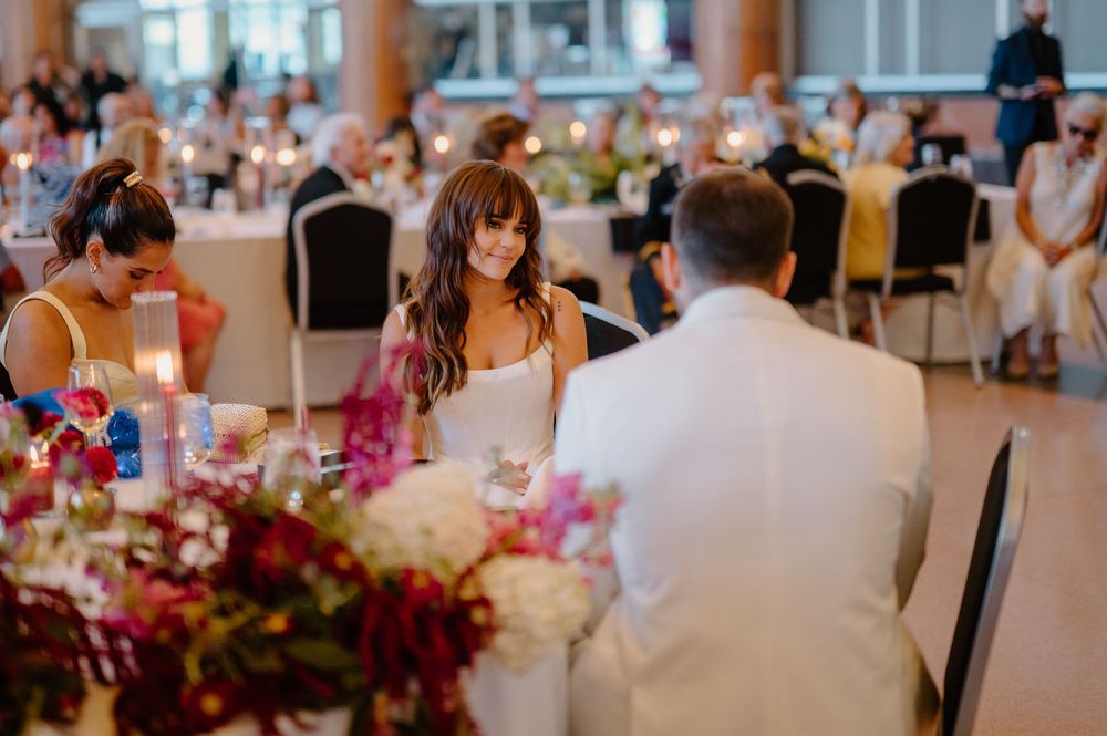 Bride smiling at guests during elegant wedding reception with floral centerpieces and candlelit tables.
