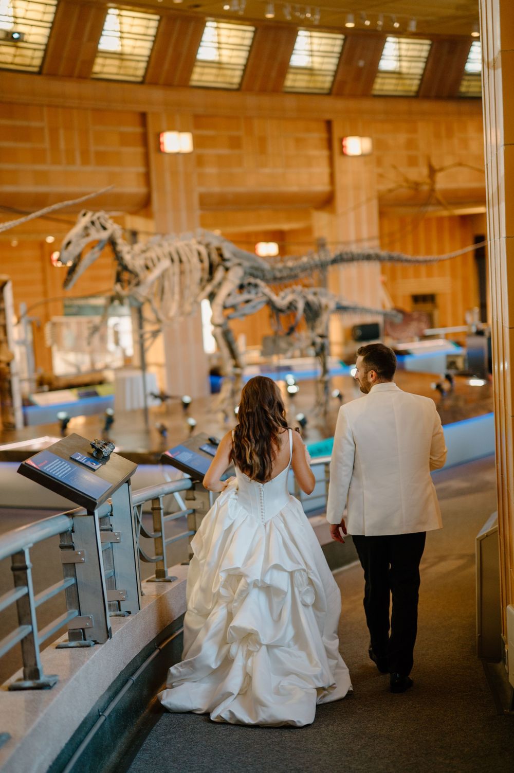 Bride and groom walk hand-in-hand through a natural history museum with dinosaur skeletons on display.