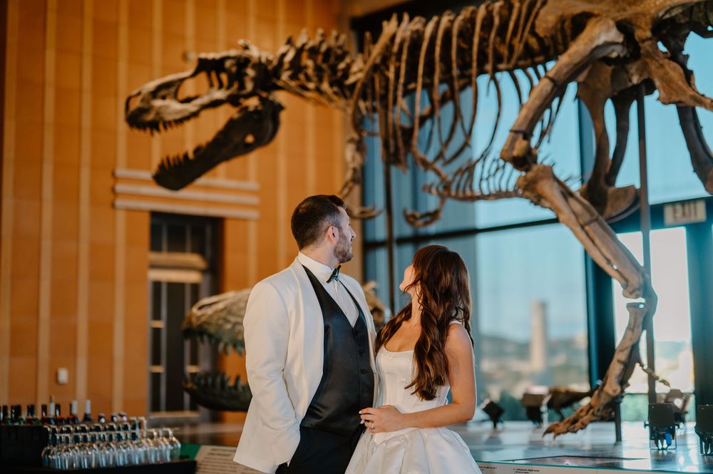 Bride and groom share romantic moment beside T-Rex skeleton at museum venue.