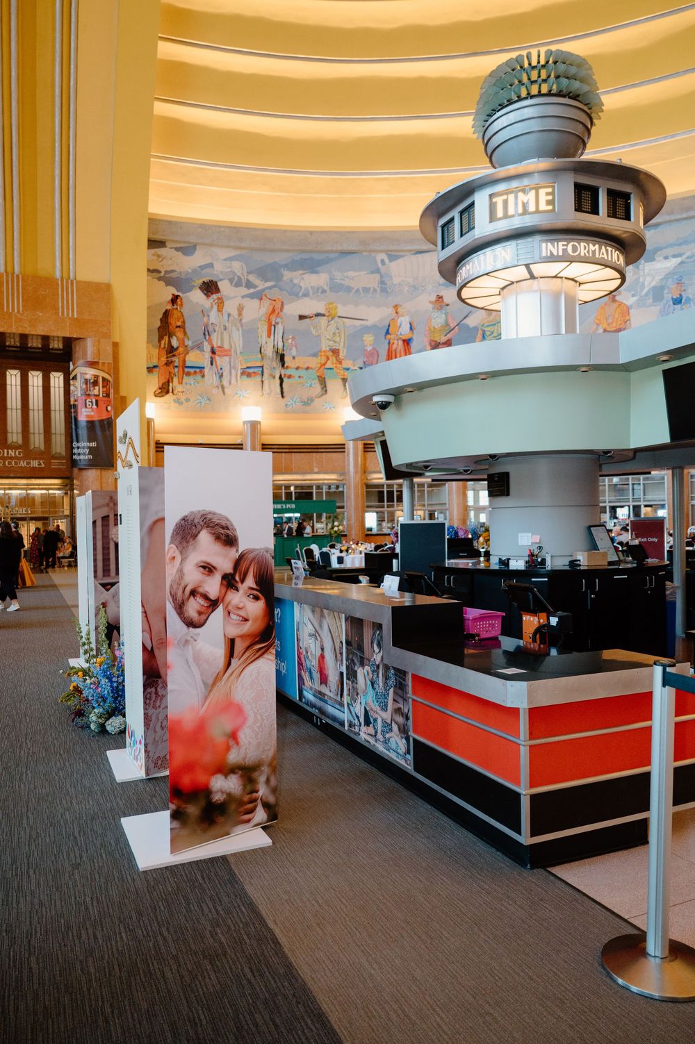 Art deco hotel lobby with information desk, large couple portrait displays, and colorful murals on curved walls.