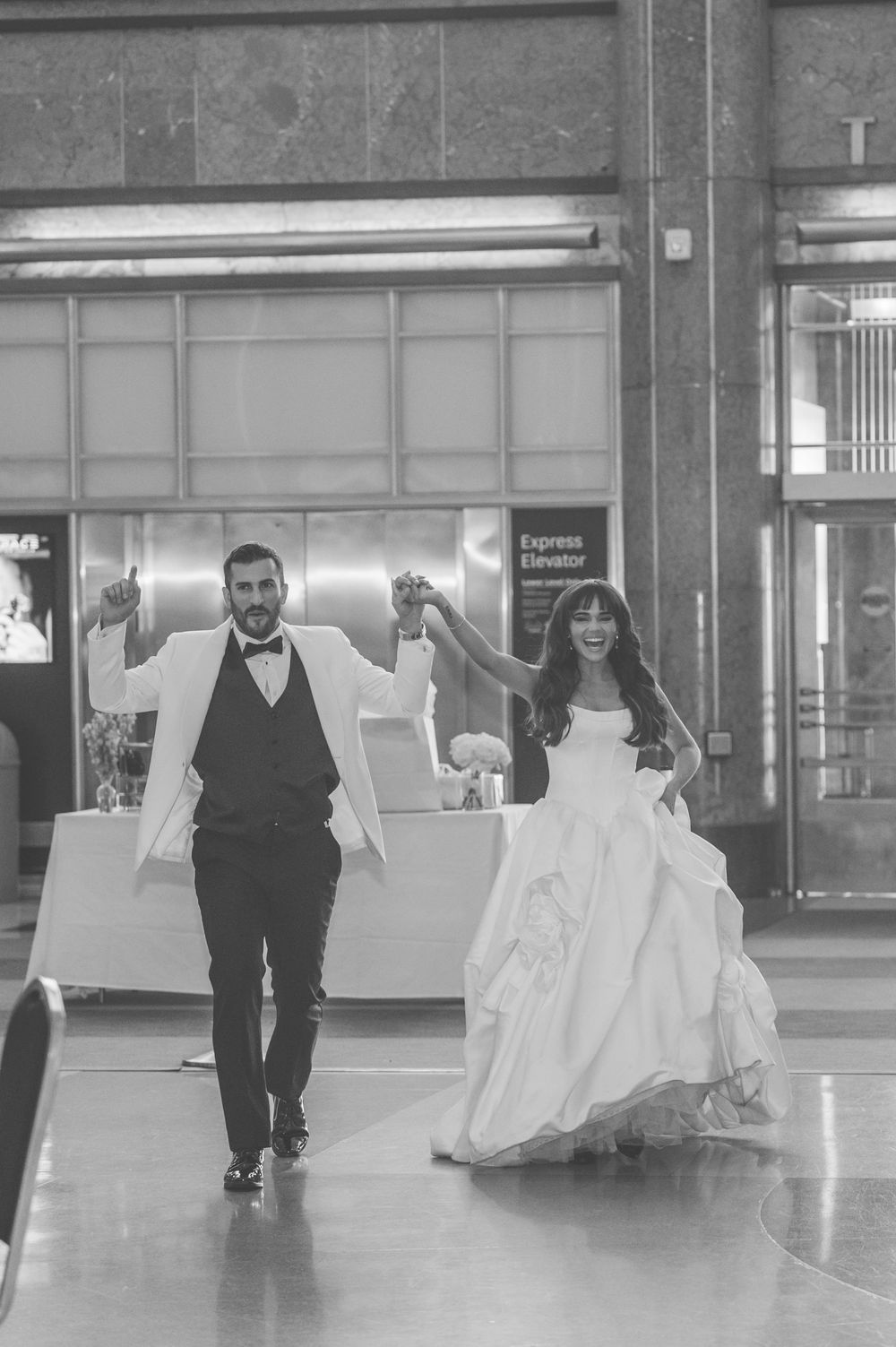 Black and white photo of a bride and groom making a grand entrance, holding hands raised in celebration.