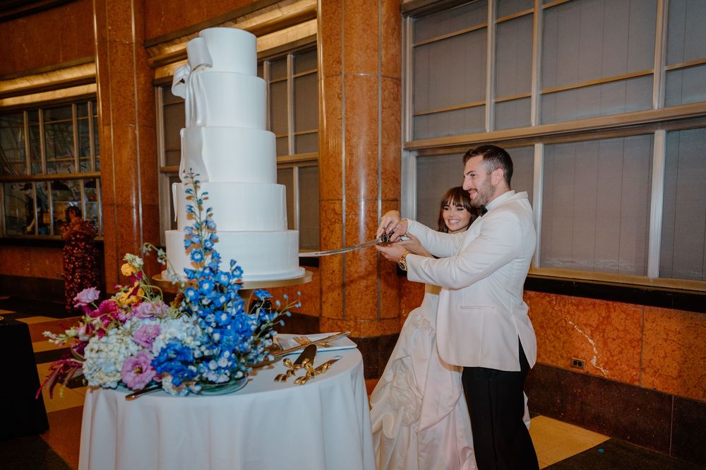 Bride and groom cutting a tall white wedding cake adorned with colorful blue and pink floral arrangements at their reception.