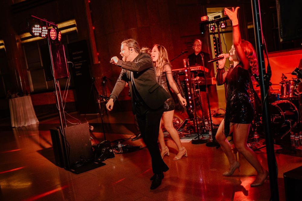 Energetic wedding band performs on stage with lead singer and dancers under dramatic red lighting.