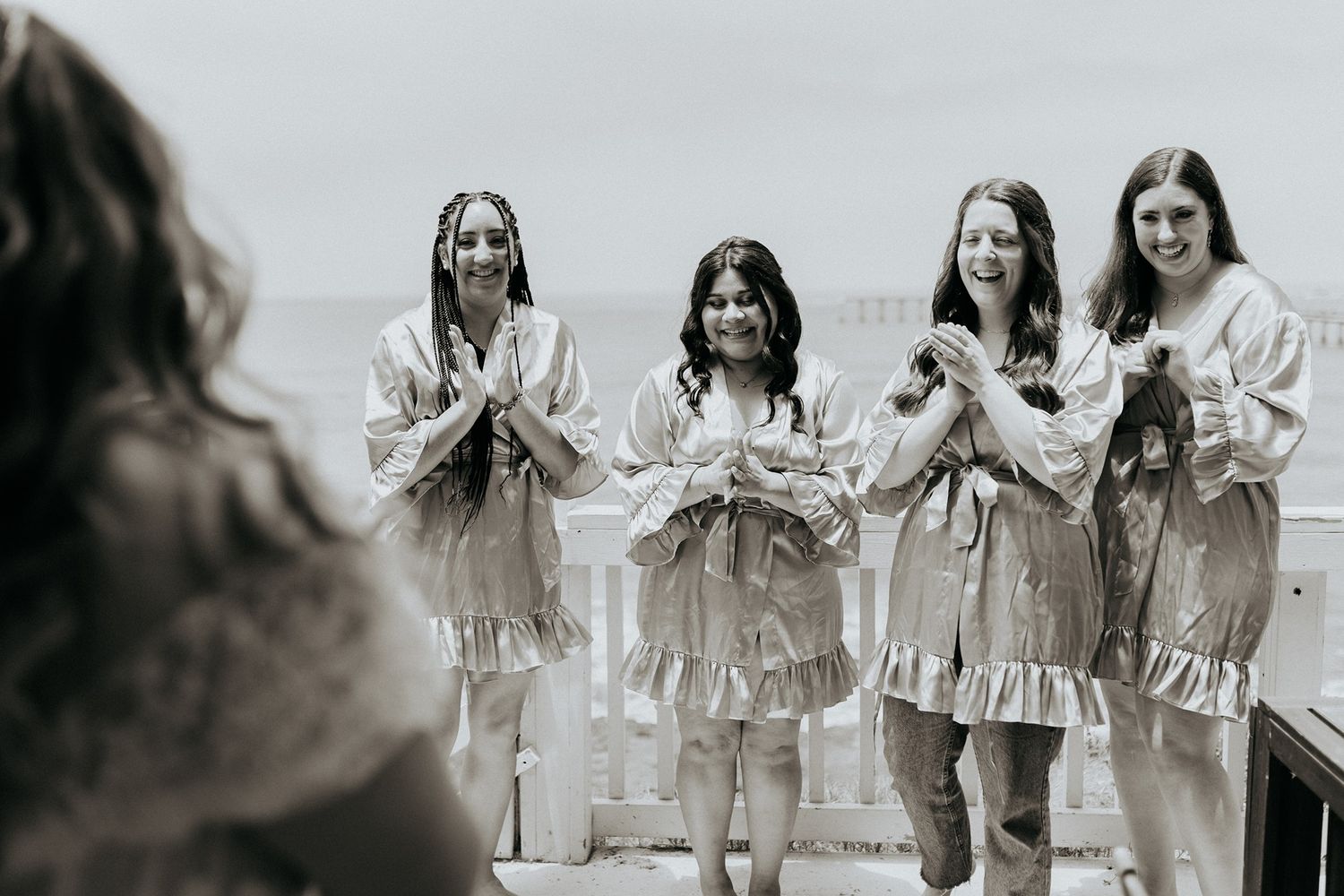 Four bridesmaids in matching robes smiling and clapping during wedding preparations in a black and white photo.