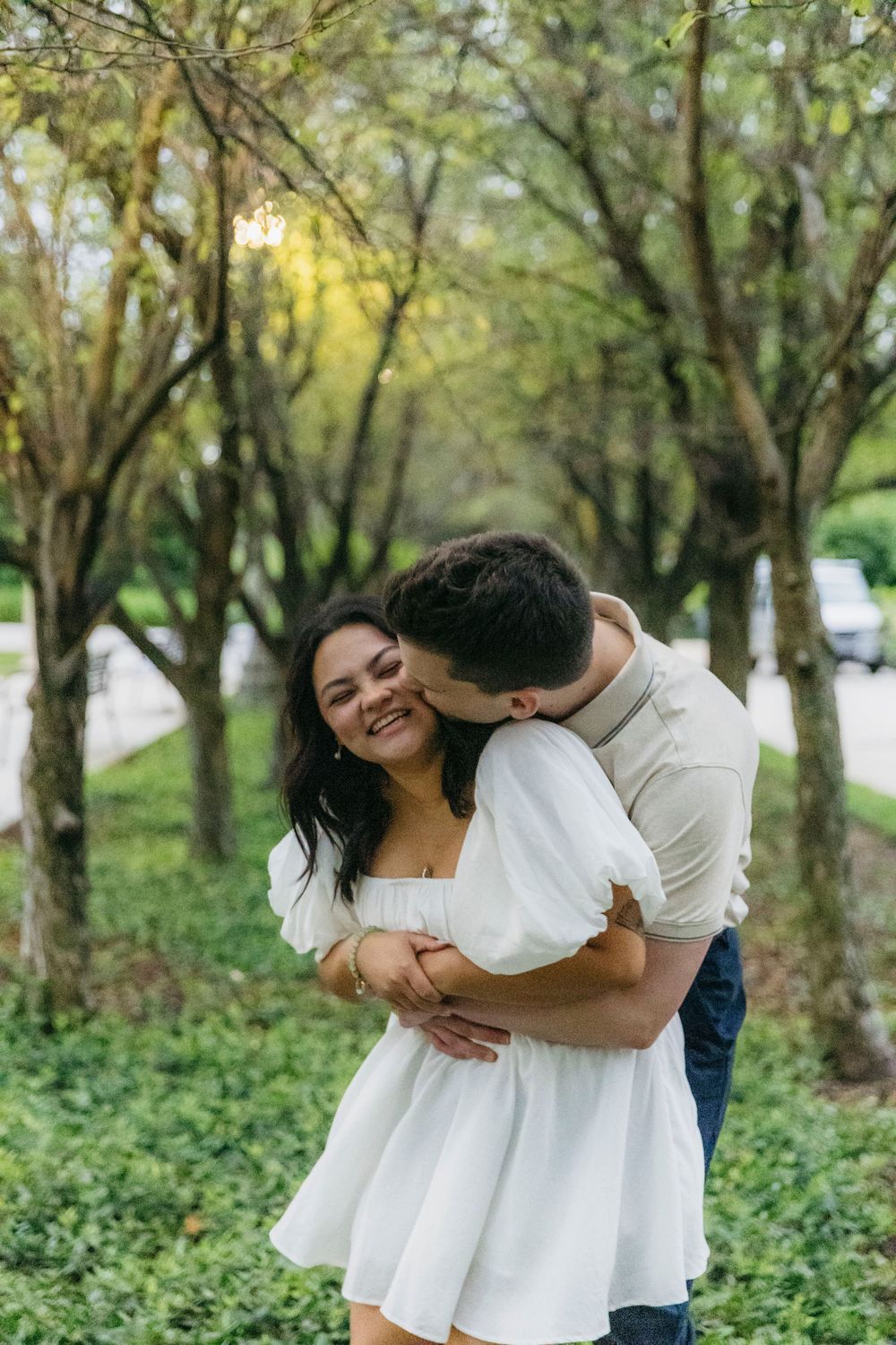 Happy couple embracing and kissing among green trees in Chicago Maggie Daley Park, woman in white mini dress smiling joyfully outdoors.